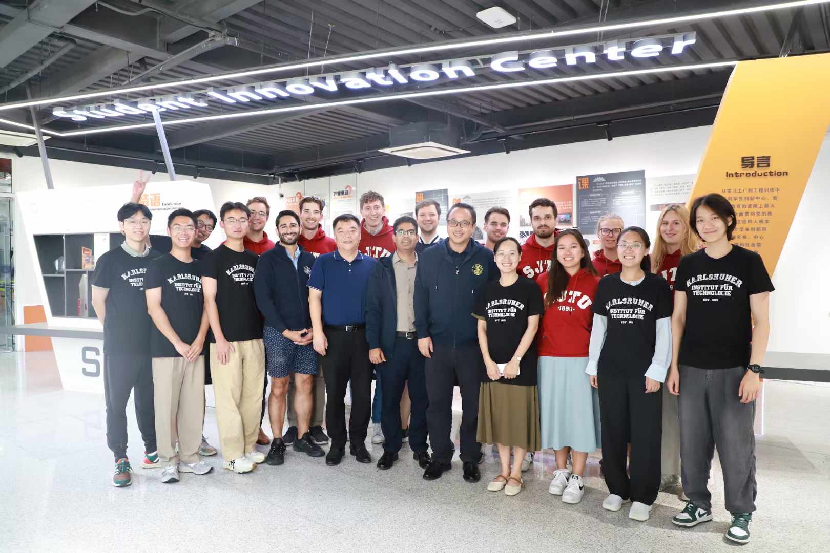 Gruppe von Menschen steht zusammen in einer Lern- oder Forschungsanlage, einige tragen Kfz- oder Uni-Shirts; ein Gel&auml;nder und Infotafeln im Hintergrund.